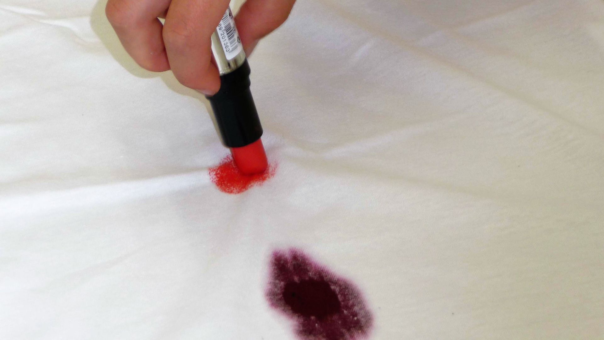 Рука пишет на белой ткани помадой рядом с пятном от красного вина