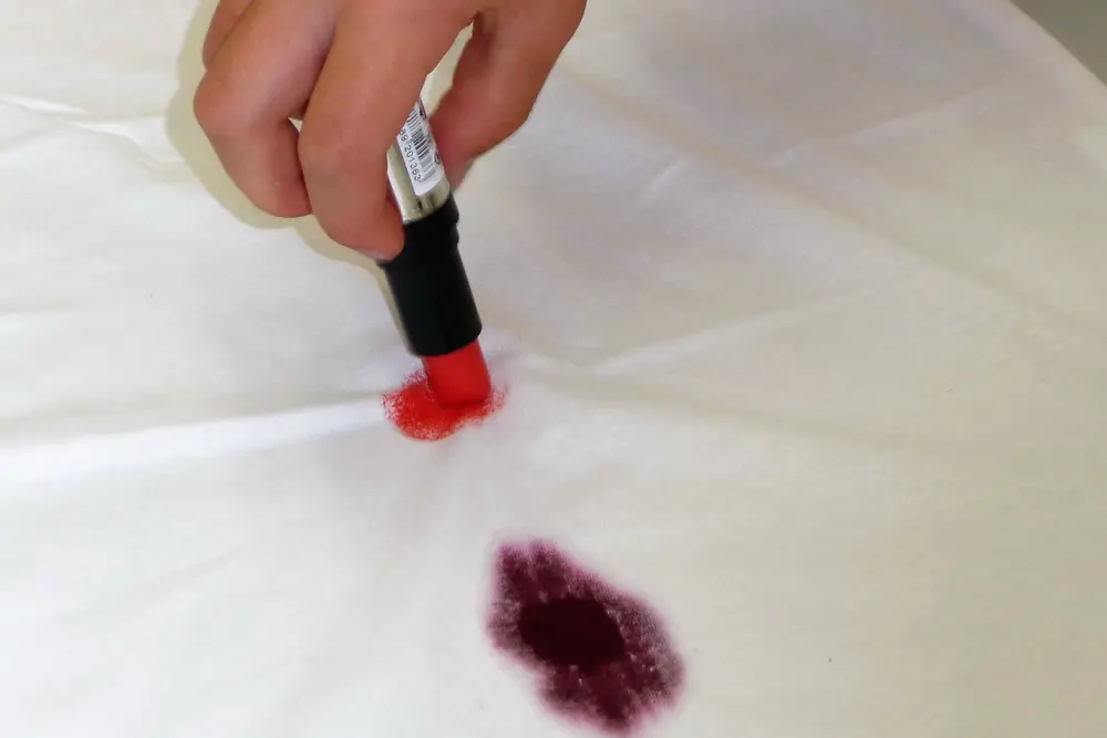 Escritura a mano sobre una tela blanca con un lápiz labial junto a una mancha de vino tinto