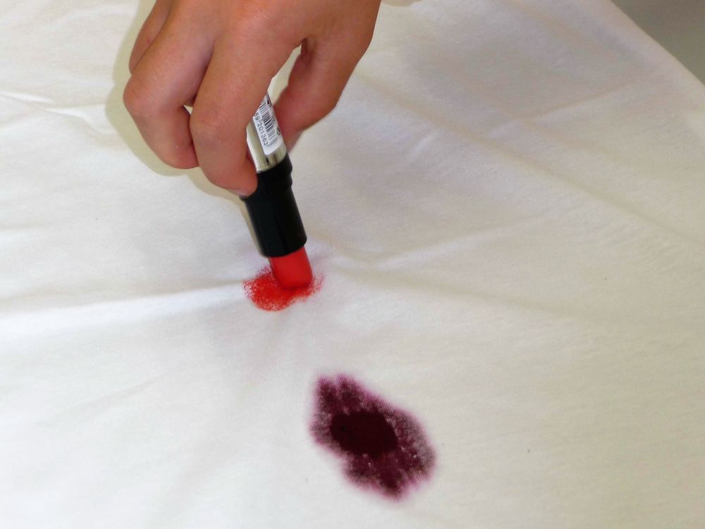 Рука пишет на белой ткани помадой рядом с пятном от красного вина