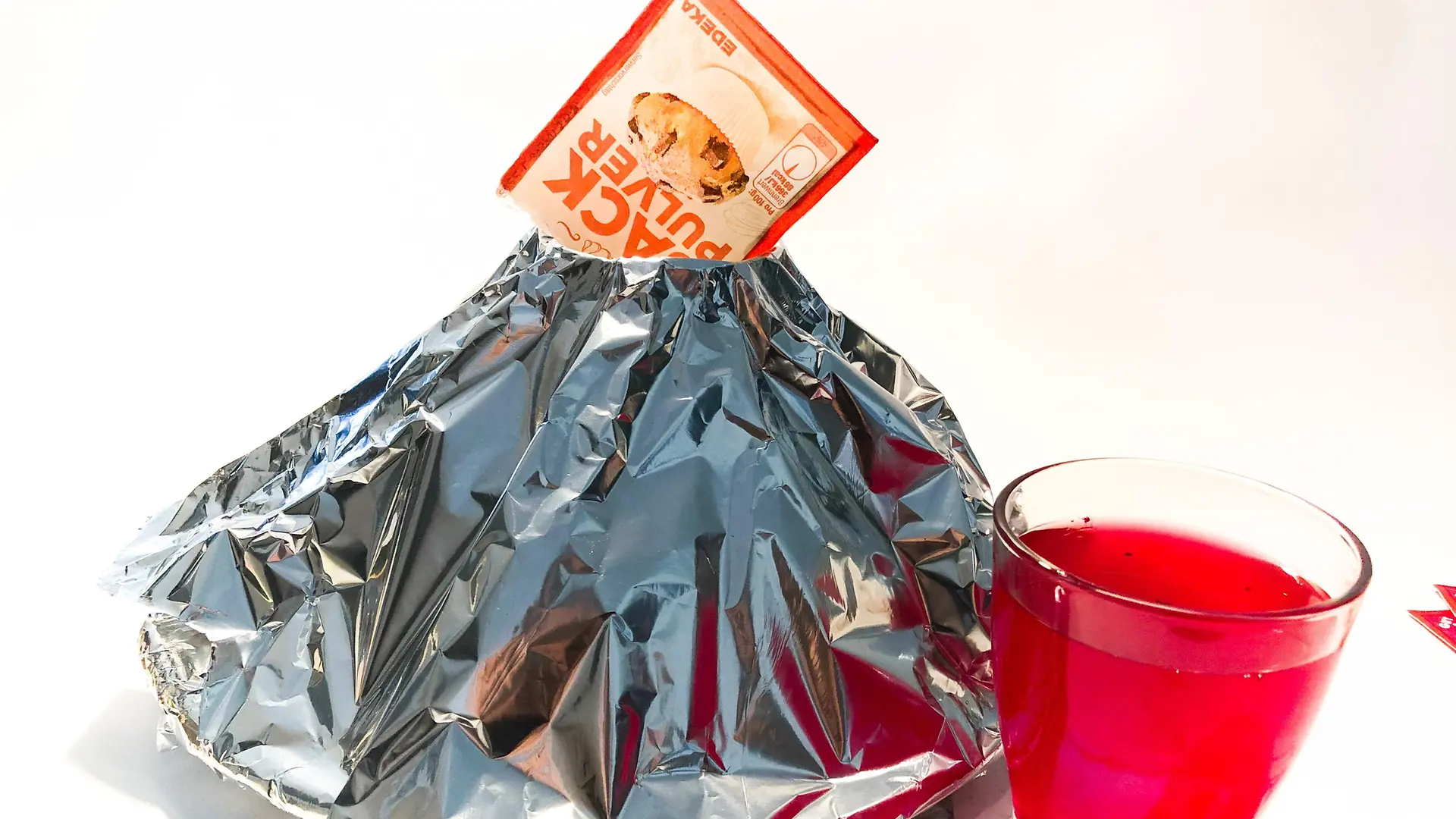 cone de papel alumínio com pacote de fermento em pó em cima e copo cheio de água vermelha