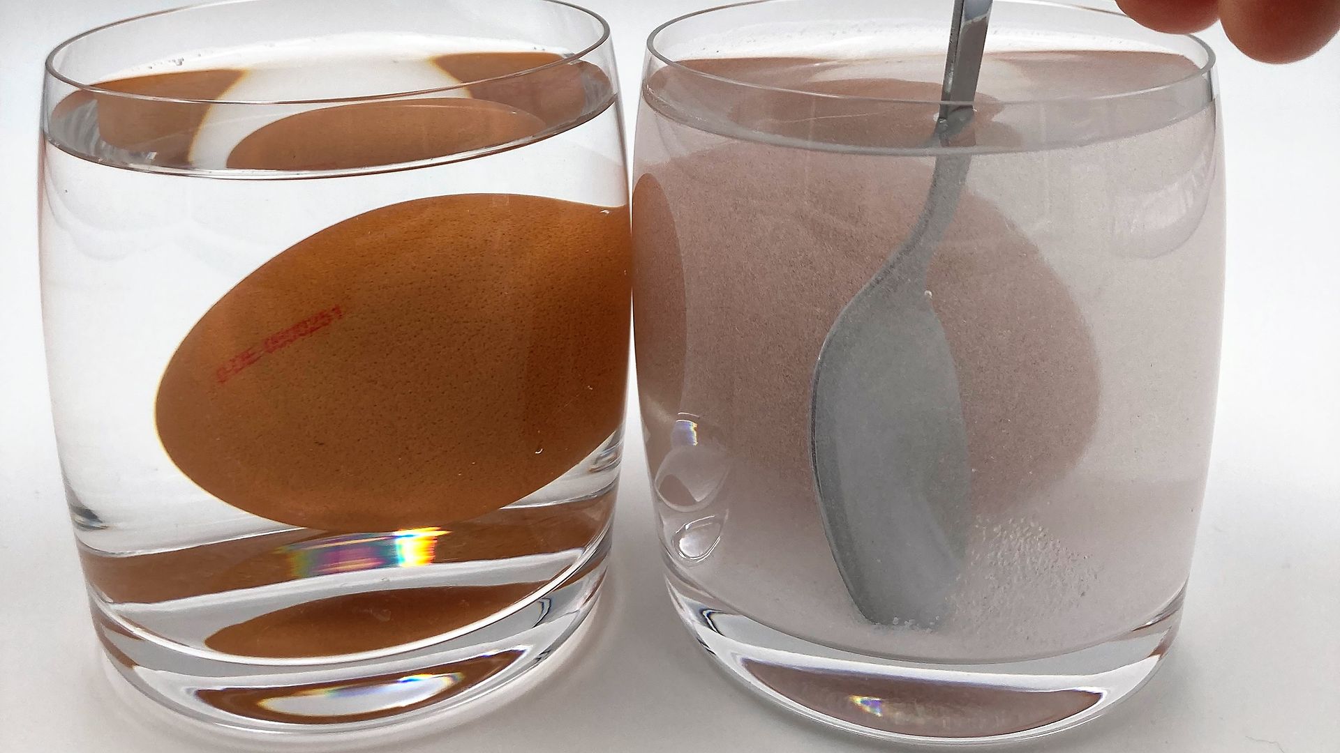 Two glasses, each filled with water and an egg. On the right side is a part of a hand stirring the salt added to the right glass with a teaspoon.