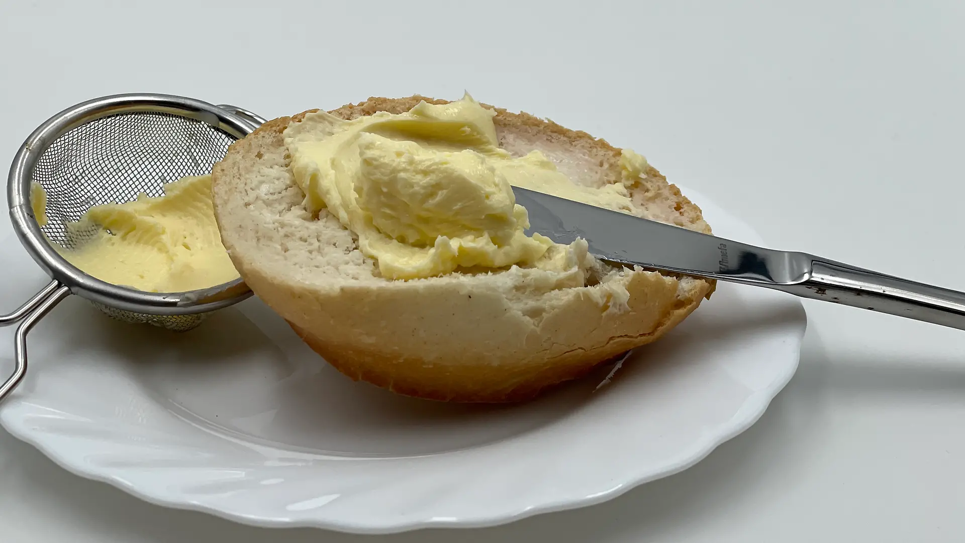 Ein halbes Brötchen mit Messer und Butter auf einem weißen Teller neben einem kleinen Sieb mit Butter