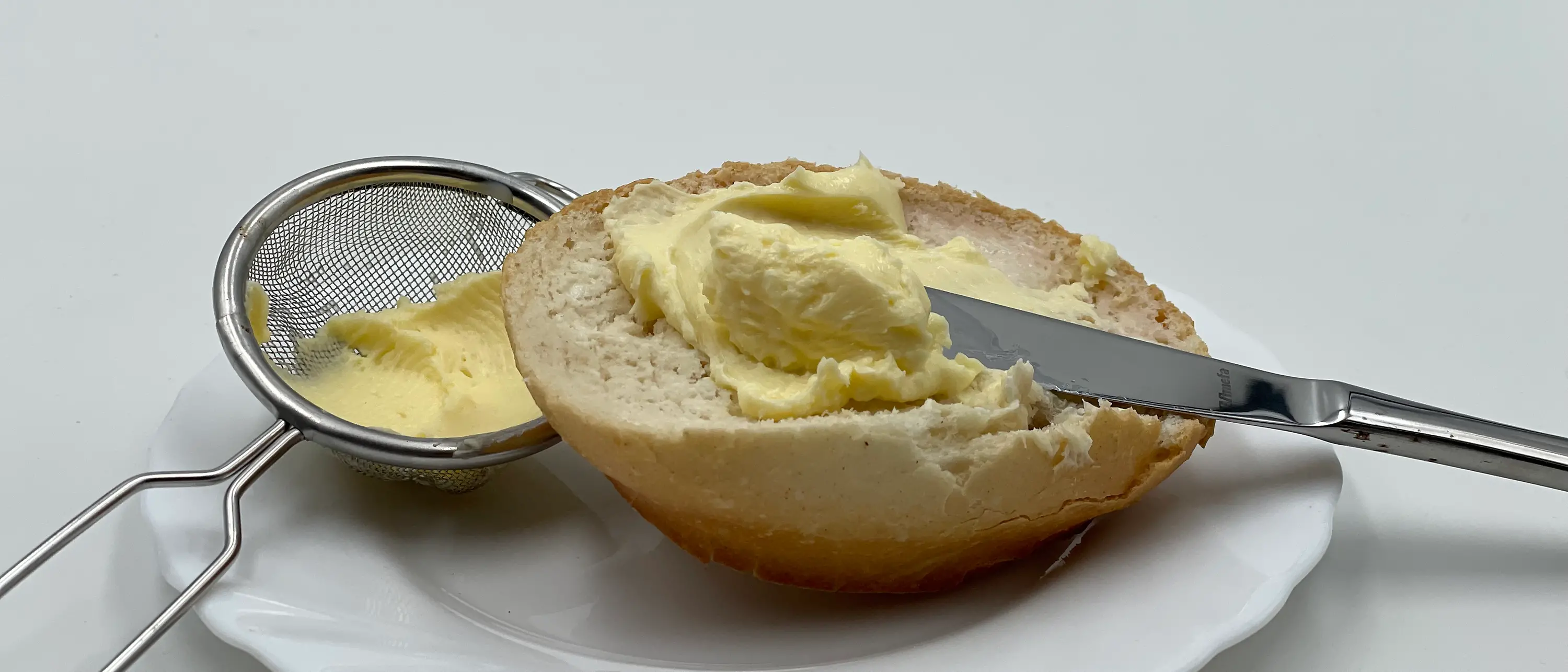 Buttered bun with knife on a plate next to a sieve with butter