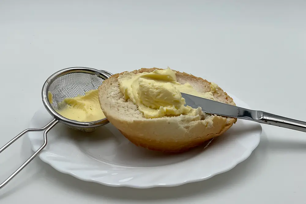 Ein halbes Brötchen mit Messer und Butter auf einem weißen Teller neben einem kleinen Sieb mit Butter