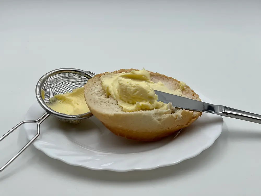 Ein halbes Brötchen mit Messer und Butter auf einem weißen Teller neben einem kleinen Sieb mit Butter