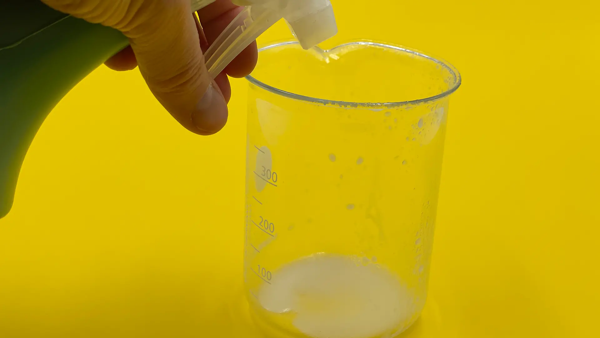 Sprühflasche, deren Inhalt in Becherglas mit weißer, schaumiger Flüssigkeit gesprüht wird.