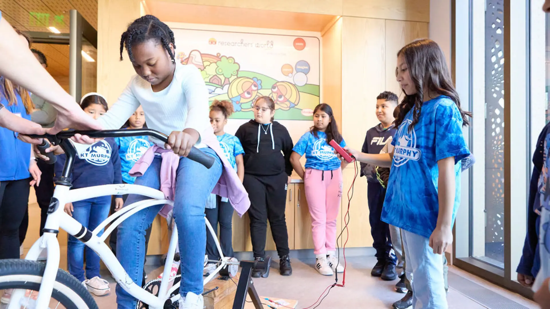 A student pedals a bike while another student operates equipment to demonstrate energy generation through motion.