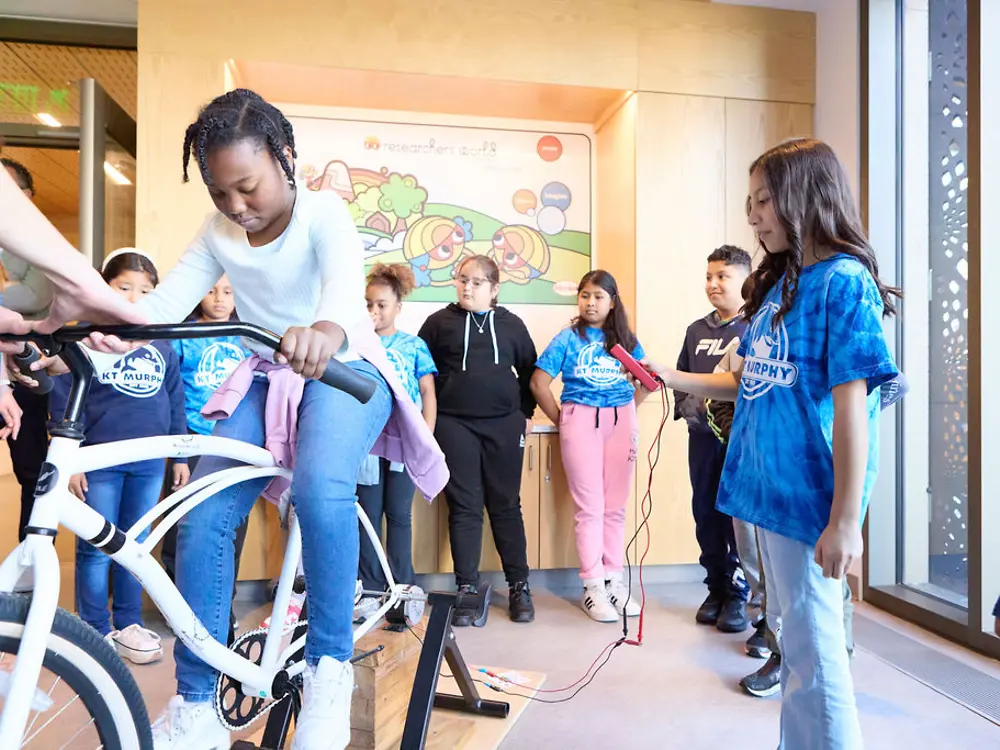 A student pedals a bike while another student operates equipment to demonstrate energy generation through motion.