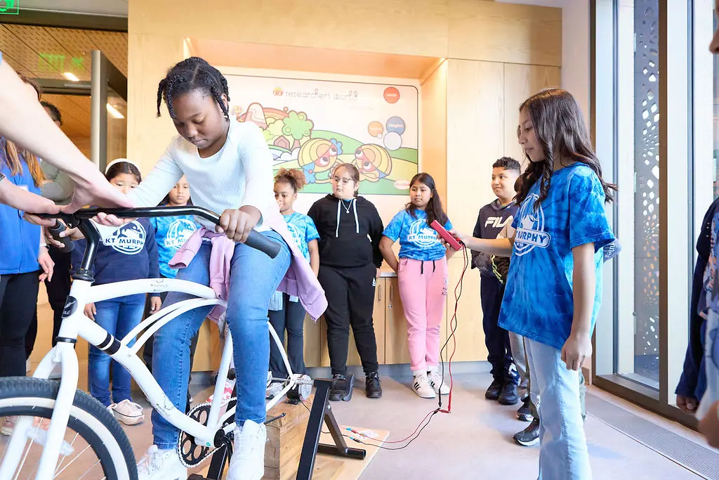 A student pedals a bike while another student operates equipment to demonstrate energy generation through motion.