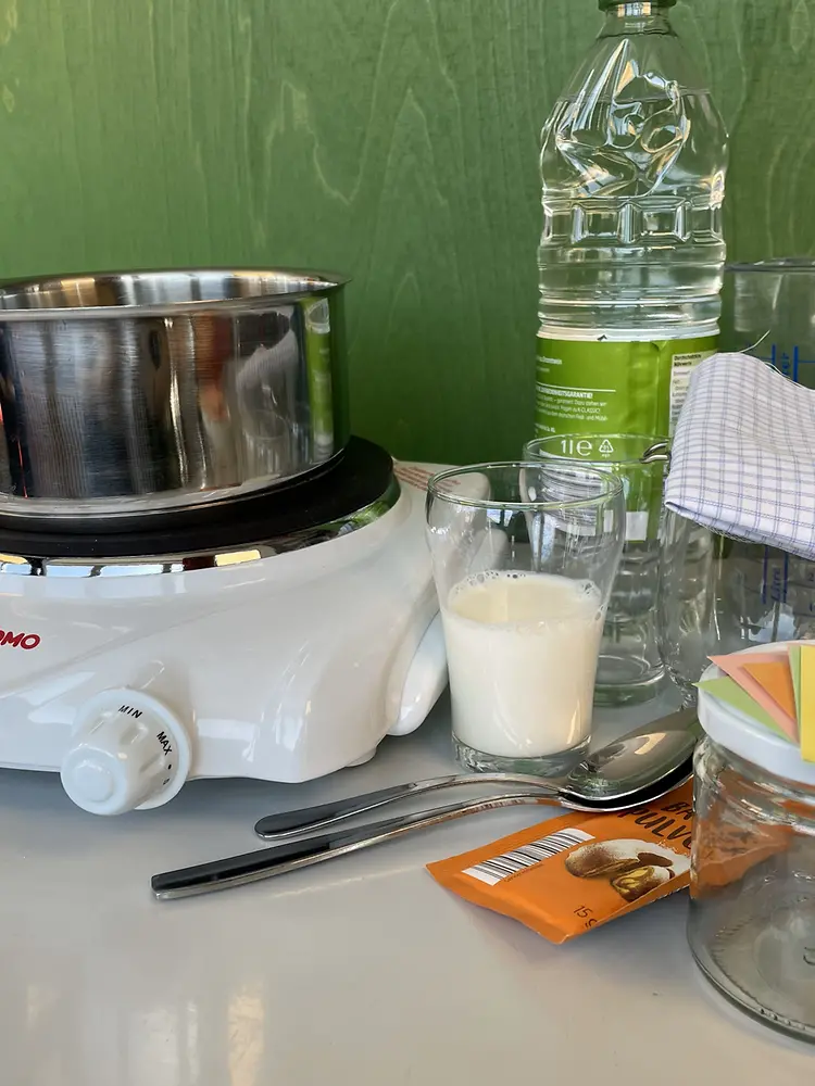 Gläser, ein Glas Kuhmilch, ein Kochtopf, ein paar Löffel, ein Trichter, eine Heizplatte, ein Messbecher, Stoffstück, ein Wasserglas, Essig, Backpulver, Papierschnipsel und ein Marmeladenglas