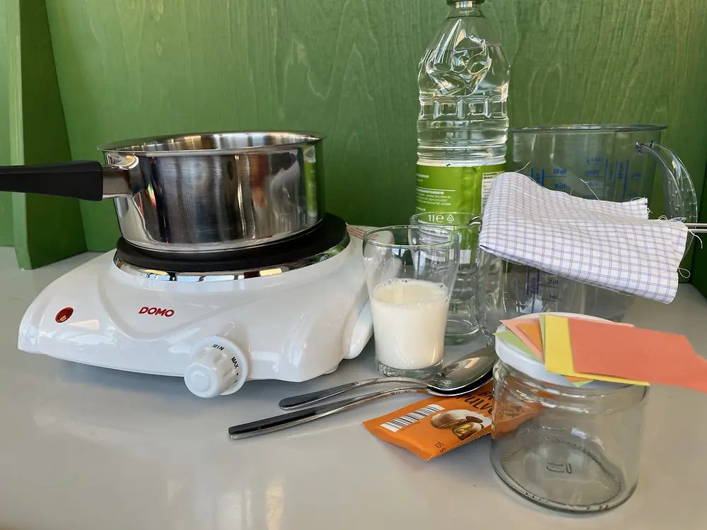 Gläser, ein Glas Kuhmilch, ein Kochtopf, ein paar Löffel, ein Trichter, eine Heizplatte, ein Messbecher, Stoffstück, ein Wasserglas, Essig, Backpulver, Papierschnipsel und ein Marmeladenglas