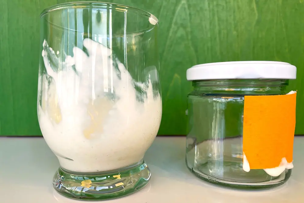 Links steht ein Wasserglas mit einer weißen cremigen Masse neben einem kleineren Glasgefäß mit weißem Schraubdeckel, auf das ein oranges Stück Papier geklebt ist. Die Gläser stehen vor einem grünen Hintergrund.