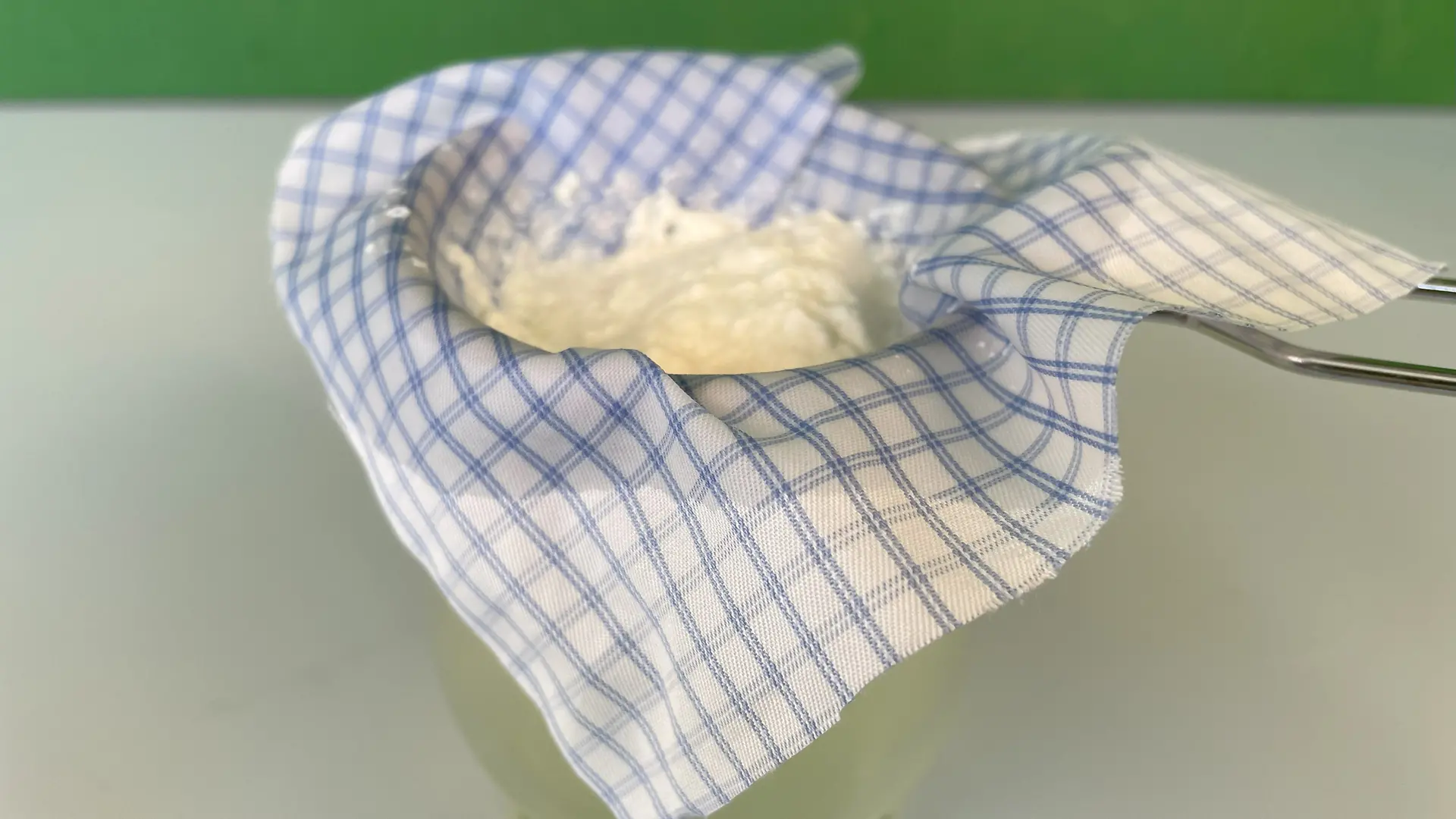 Glas, auf dem ein Sieb mit einem Stoffstück liegt. Auf dem Stoffstück ist die geronnene Milch zu sehen.