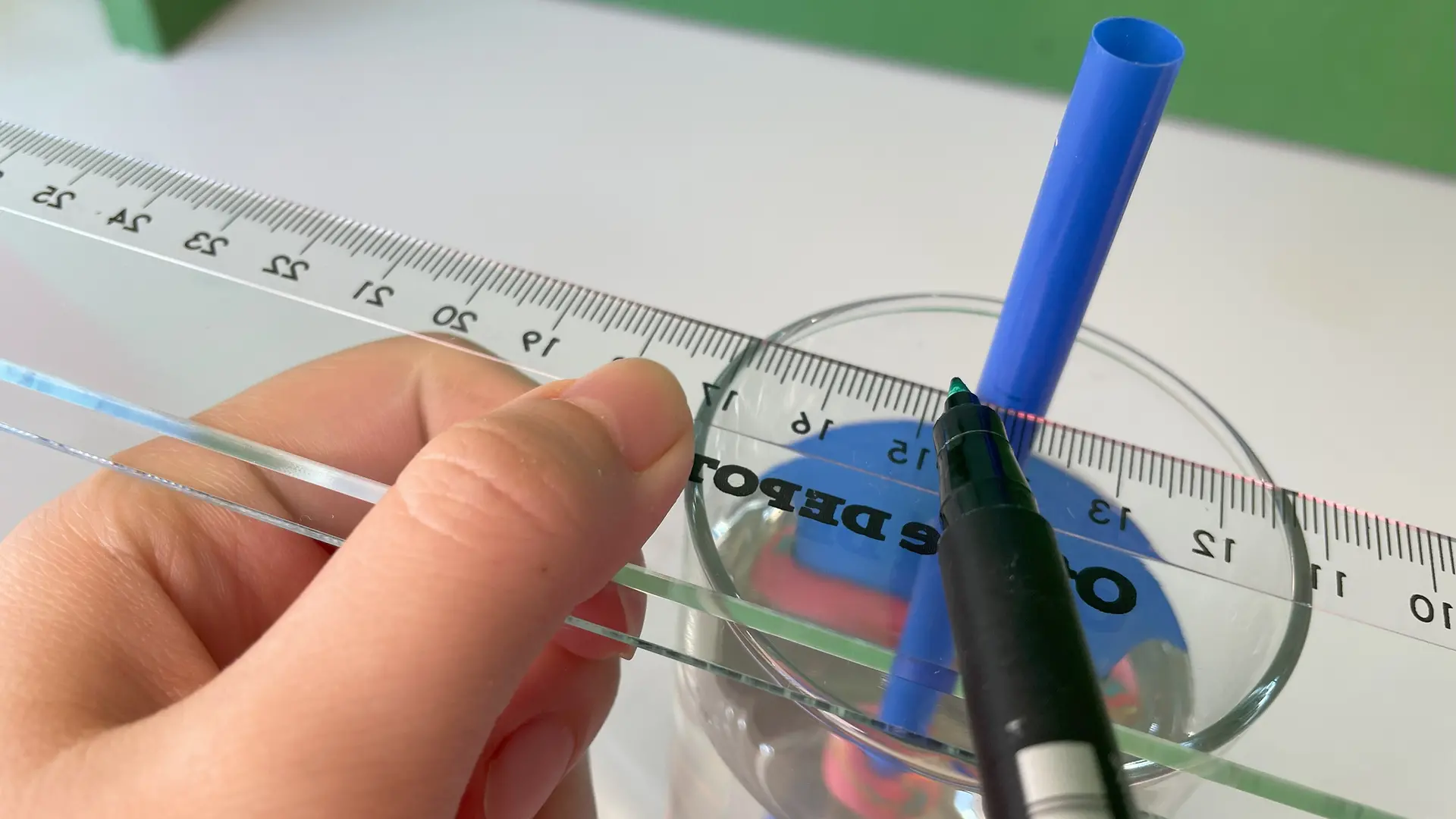 Nahaufnahme: Ein Lineal liegt auf einem Glas und berührt einen blauen Strohhalm. An der Stelle, wo das Lineal den Strohhalm berührt, wird mit einem schwarzen Filzstift eine Markierung auf dem Strohhalm angebracht.