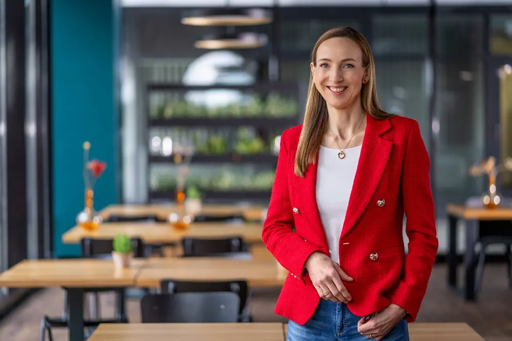 Dr. Simone Bagel-Trah trägt einen roten Blazer und ein weißes Top in einer modernen Umgebung mit Holztischen und Pflanzen im Hintergrund.
