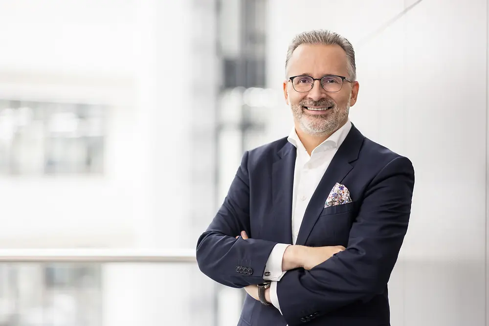 Carsten Knobel trägt einen Anzug und steht mit verschränkten Armen in einer hellen, modernen Büroumgebung. 