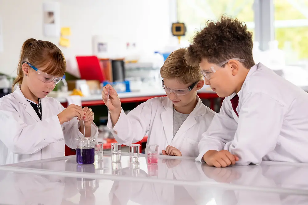 Drei Kinder in Laborkitteln und Schutzbrillen führen gemeinsam ein Experiment mit farbigen Flüssigkeiten durch.